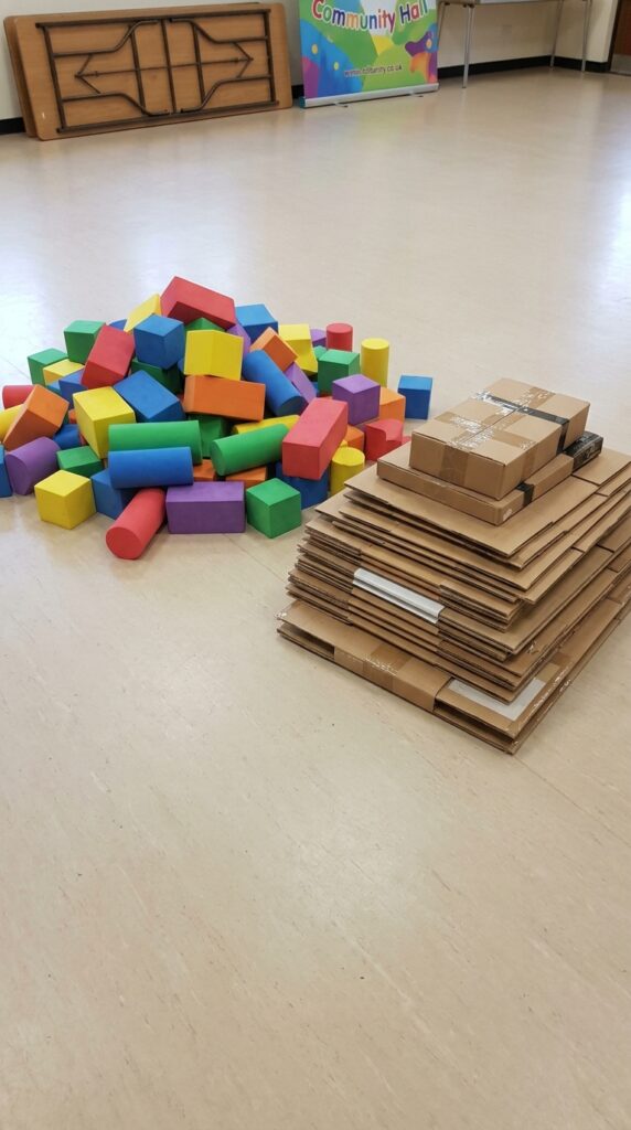 A play area is set up with a table or floor space containing neatly arranged foam blocks or cardboard boxes in separate piles. A marked build zone using tape or cones defines the play area, where children are encouraged to construct the tallest tower within a limited time.